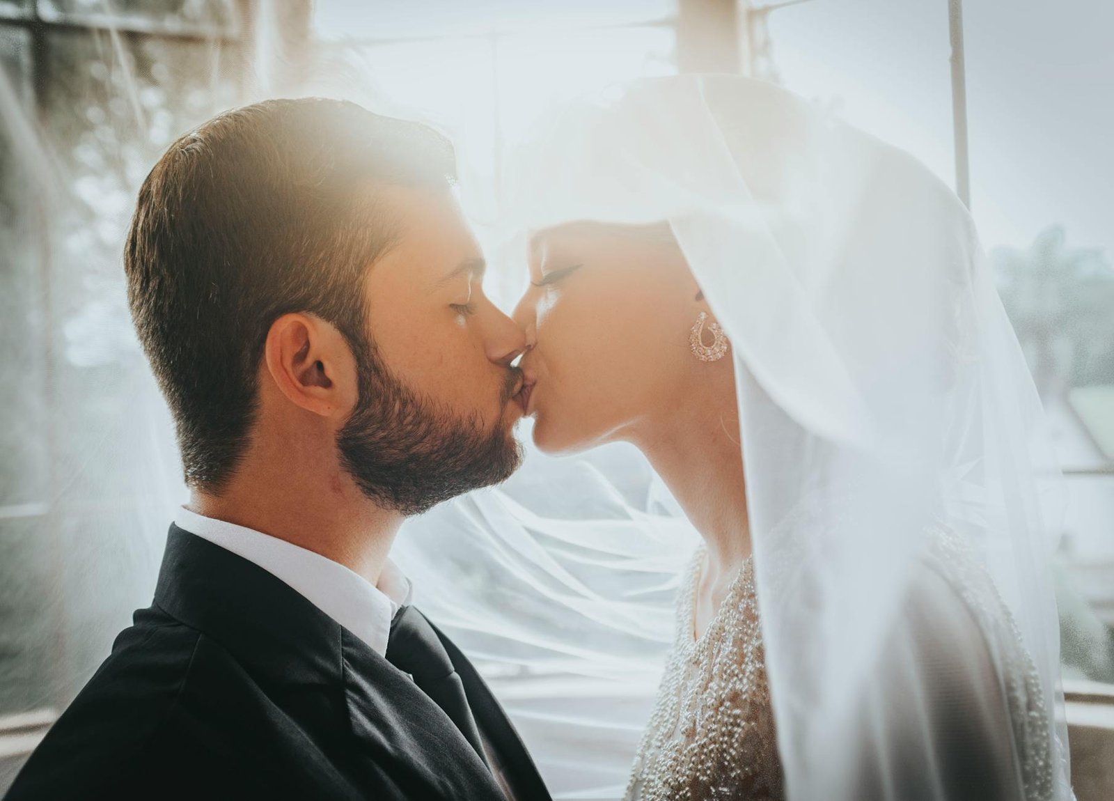 A bride and groom share a romantic kiss in elegant wedding attire, indoors with soft lighting.